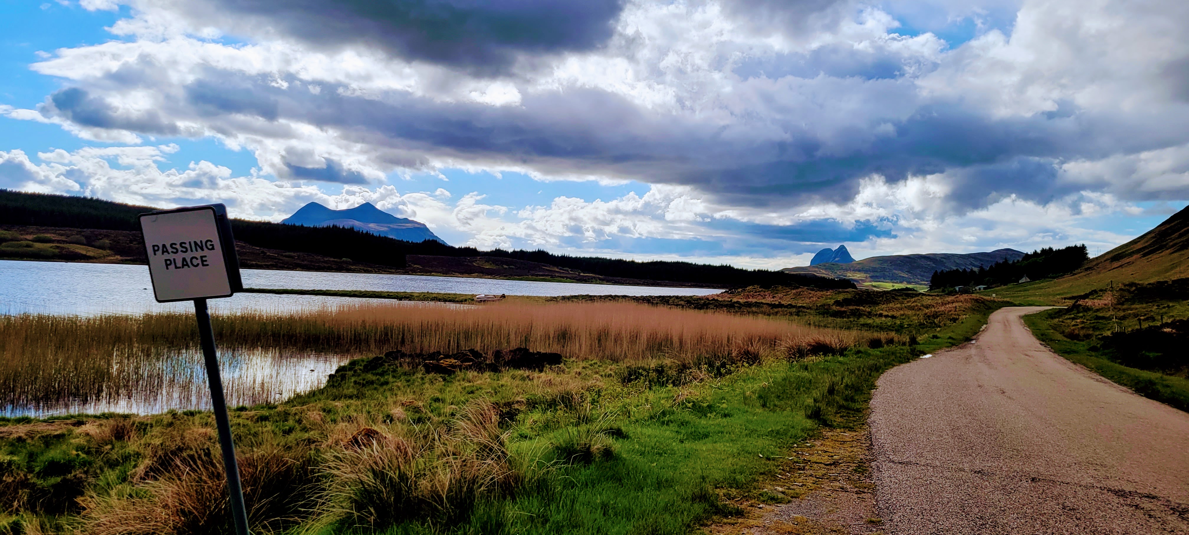 single lane road next to a lake with hills and two mountains in the distance with a sign that says passing place